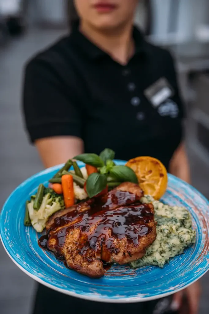 Kelner trzyma talerz z grillowanym mięsem BBQ, puree ziołowym, warzywami i cytryną.
