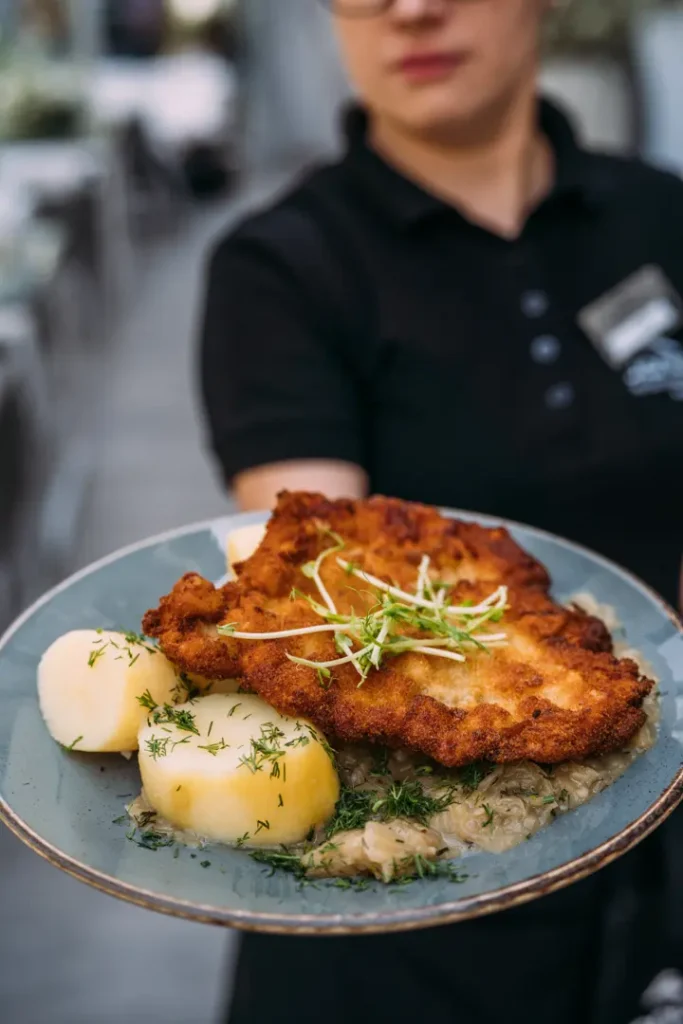 Panierowany kotlet schabowy, ziemniaki z koperkiem i kapusta z dymką na talerzu podanym przez kelnera.