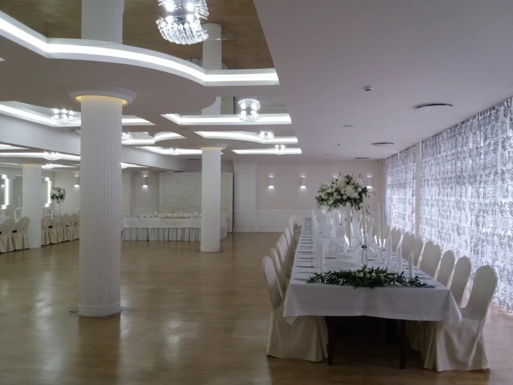 Jasna sala bankietowa z żyrandolami, białymi kolumnami i stołem, idealna na eleganckie przyjęcie.