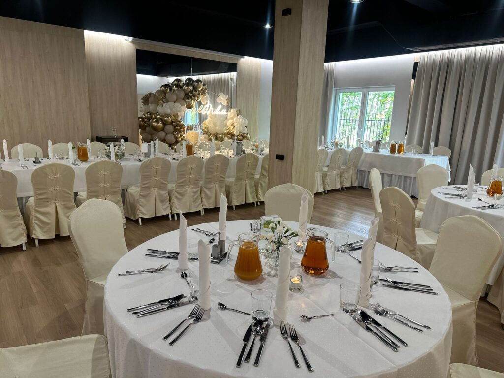 Sala bankietowa z okrągłymi stołami, białymi obrusami, ozdobnym łukiem z balonów i napisem „Mr & Mrs”.