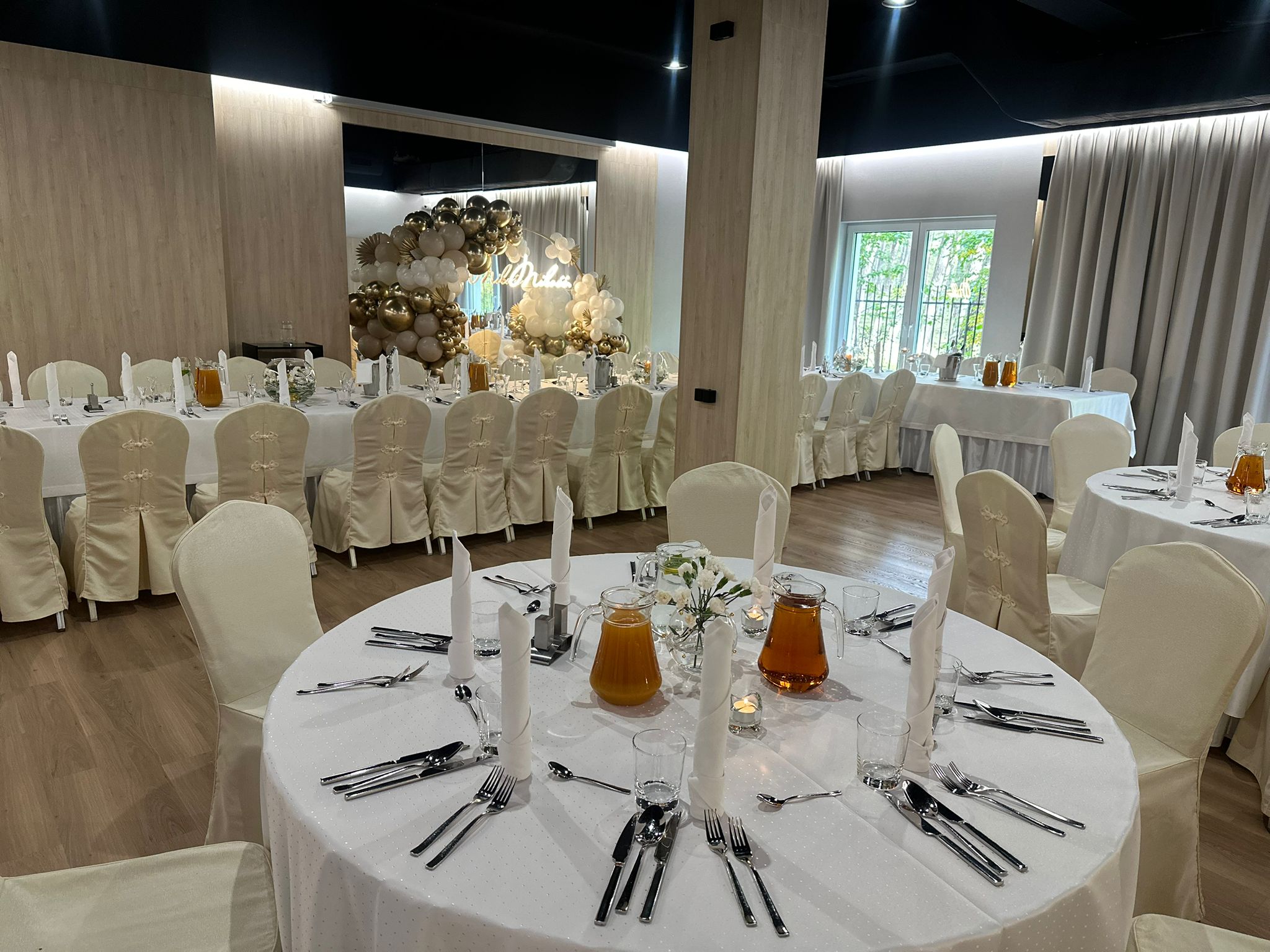 Banquet hall featuring round tables, white tablecloths, a decorative balloon arch, and a 'Mr & Mrs' sign.