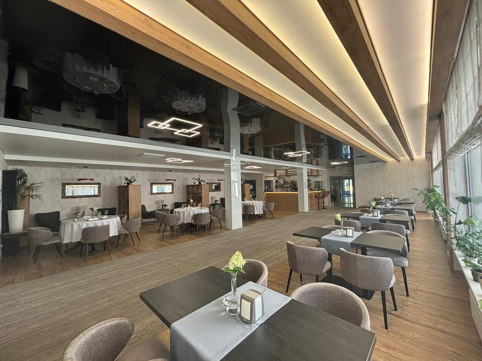Nowoczesna restauracja z drewnianą podłogą, dużymi oknami i roślinami, a stoły ozdobione są wazonami.