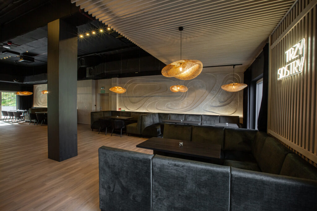 Wnętrze restauracji z zielonymi lożami, drewnianymi stolikami i wiszącymi lampami.