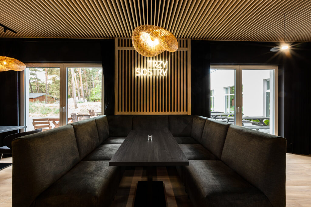 Nowoczesne wnętrze restauracji z szarą sofą w kształcie litery U, stołem i dużymi oknami. Neon „TRZY SIOSTRY”.