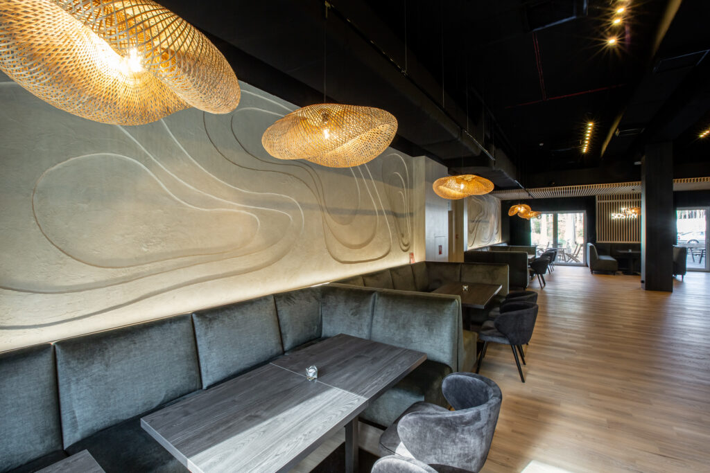 Nowoczesne wnętrze restauracji, szare welurowe loże, jasne drewniane stoły i duże okna.